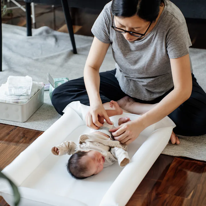 A seated parent gently dressing a newborn on a white changing pad on the floor, with diaper supplies...
