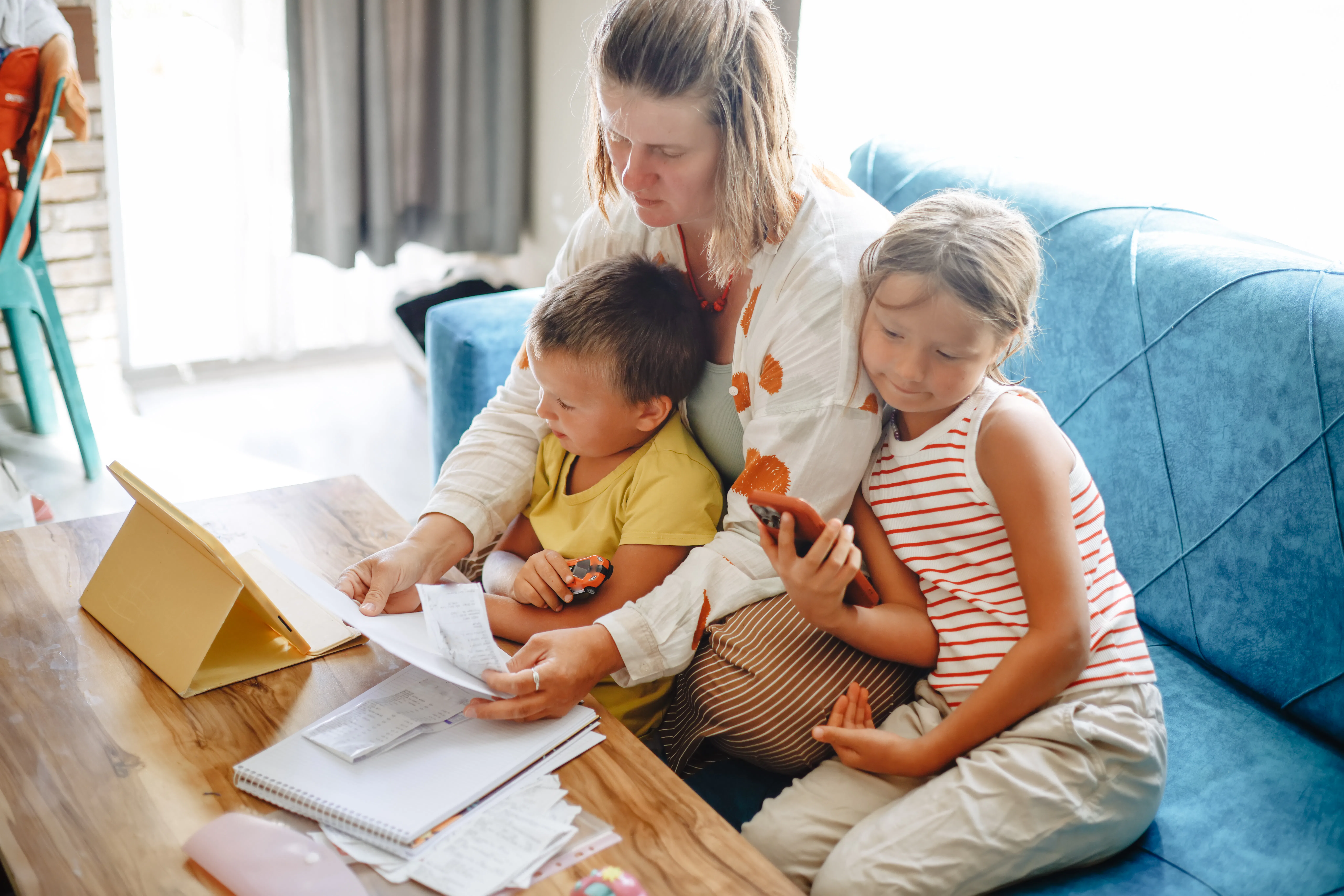Parent on a blue couch helping two children with papers and a tablet at a wooden table, surrounded b...