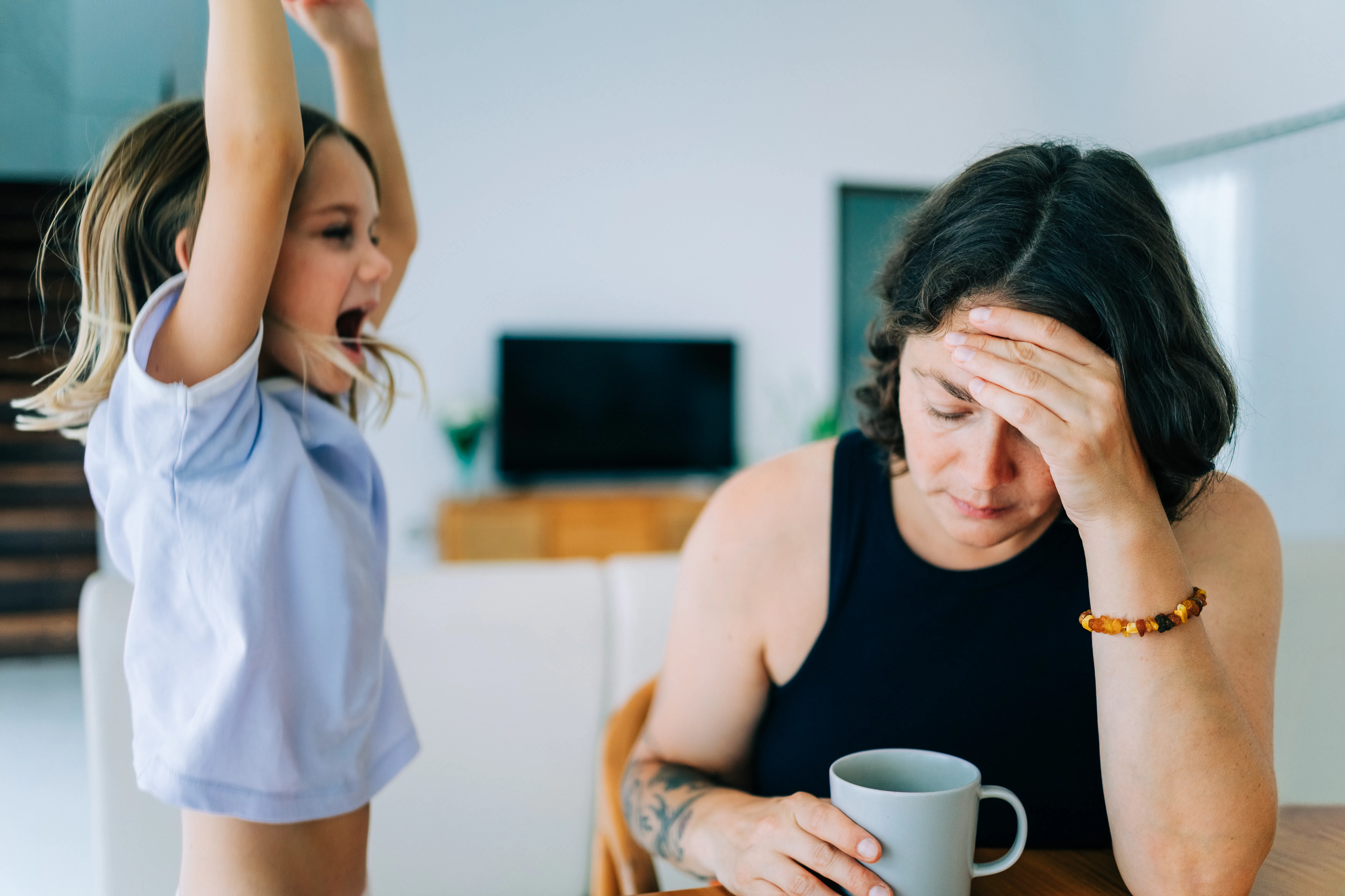 Tired adult with coffee holding head at kitchen table while energetic child stands nearby with arms ...
