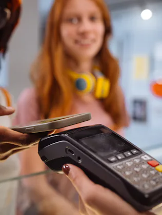Two young women at a counter; one uses a smartphone to tap-pay on a card terminal held by a cashier,...