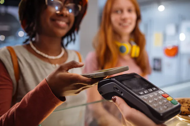 Two young women at a counter; one uses a smartphone to tap-pay on a card terminal held by a cashier,...