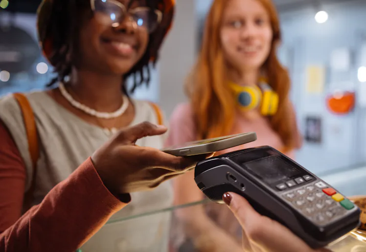 Two young women at a counter; one uses a smartphone to tap-pay on a card terminal held by a cashier,...