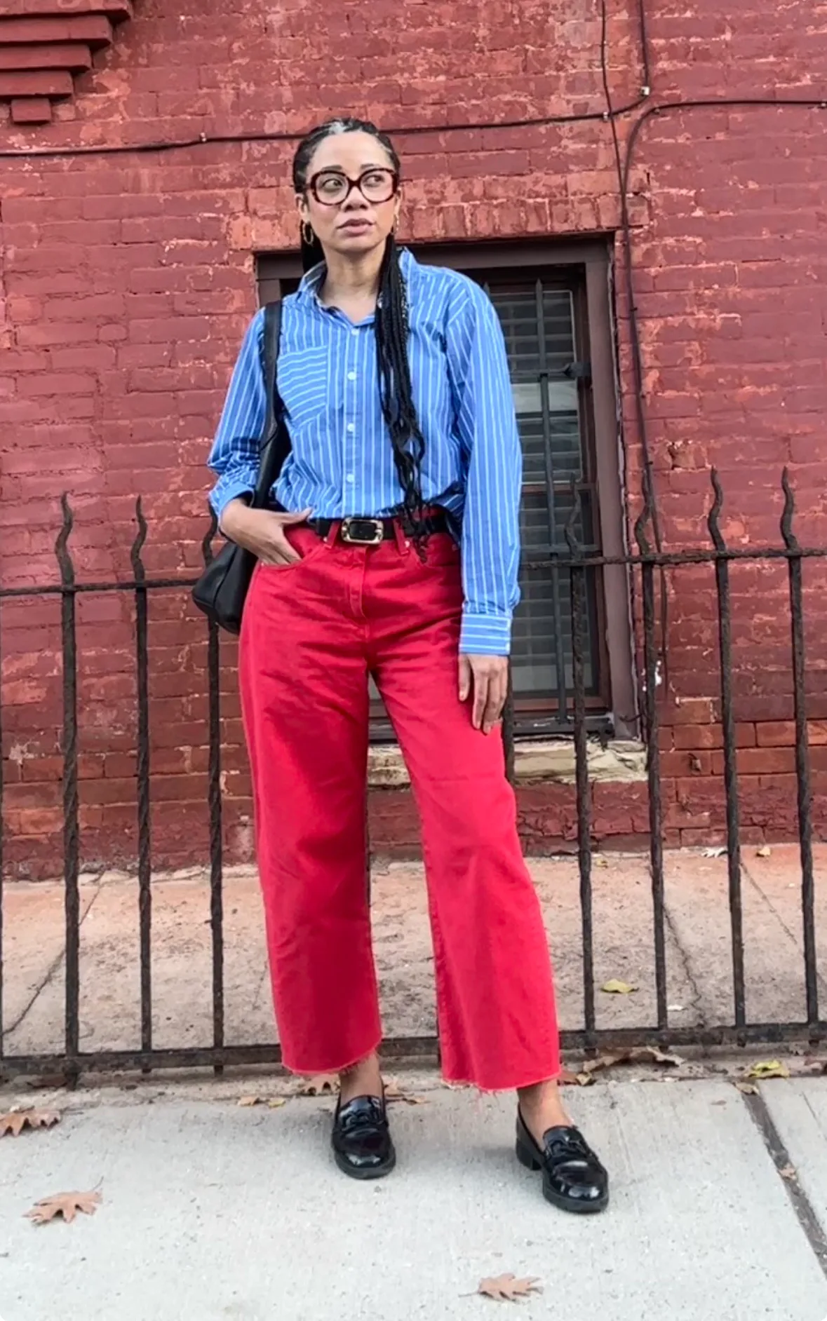 Beauty news writer Eden Stuart in red jeans with a blue button down shirt, standing in front of a br...