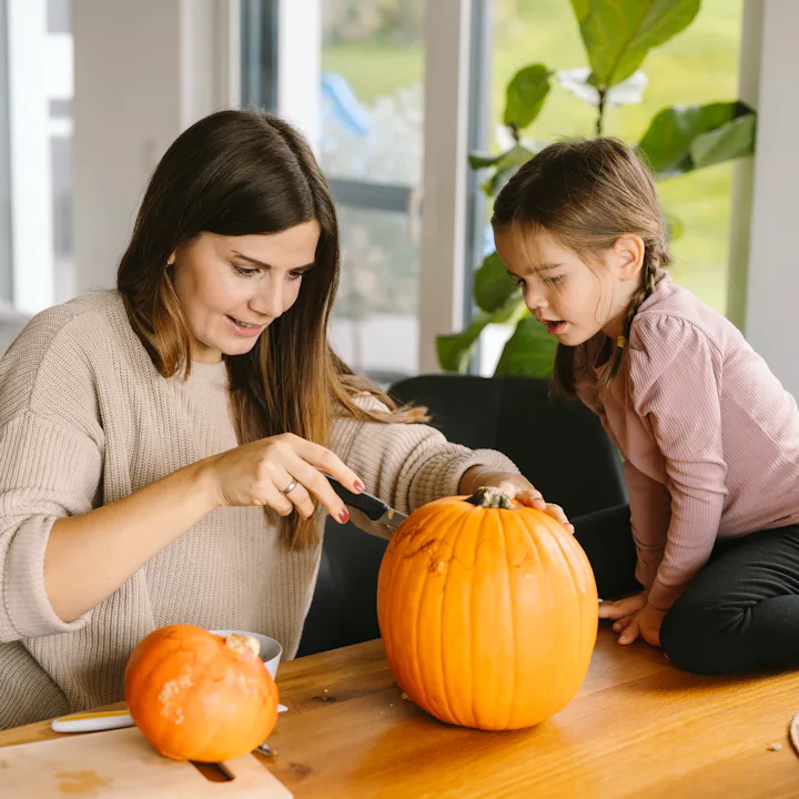 A woman carving a large pumpkin at a wooden table while a young girl watches closely, with small pum...