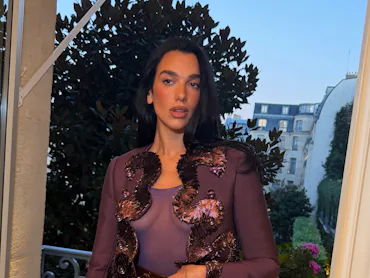 Dua Lipa standing in front of a window in Paris, wearing a purple jacket and black hot shorts
