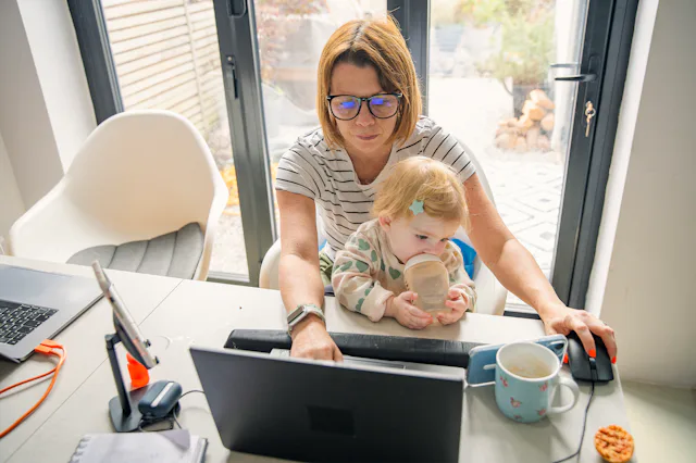 Woman wearing glasses works at a laptop in a home office, holding a toddler on her lap who drinks fr...