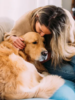 Woman sits on a couch leaning forward to kiss and pet a large golden retriever curled beside her, bo...