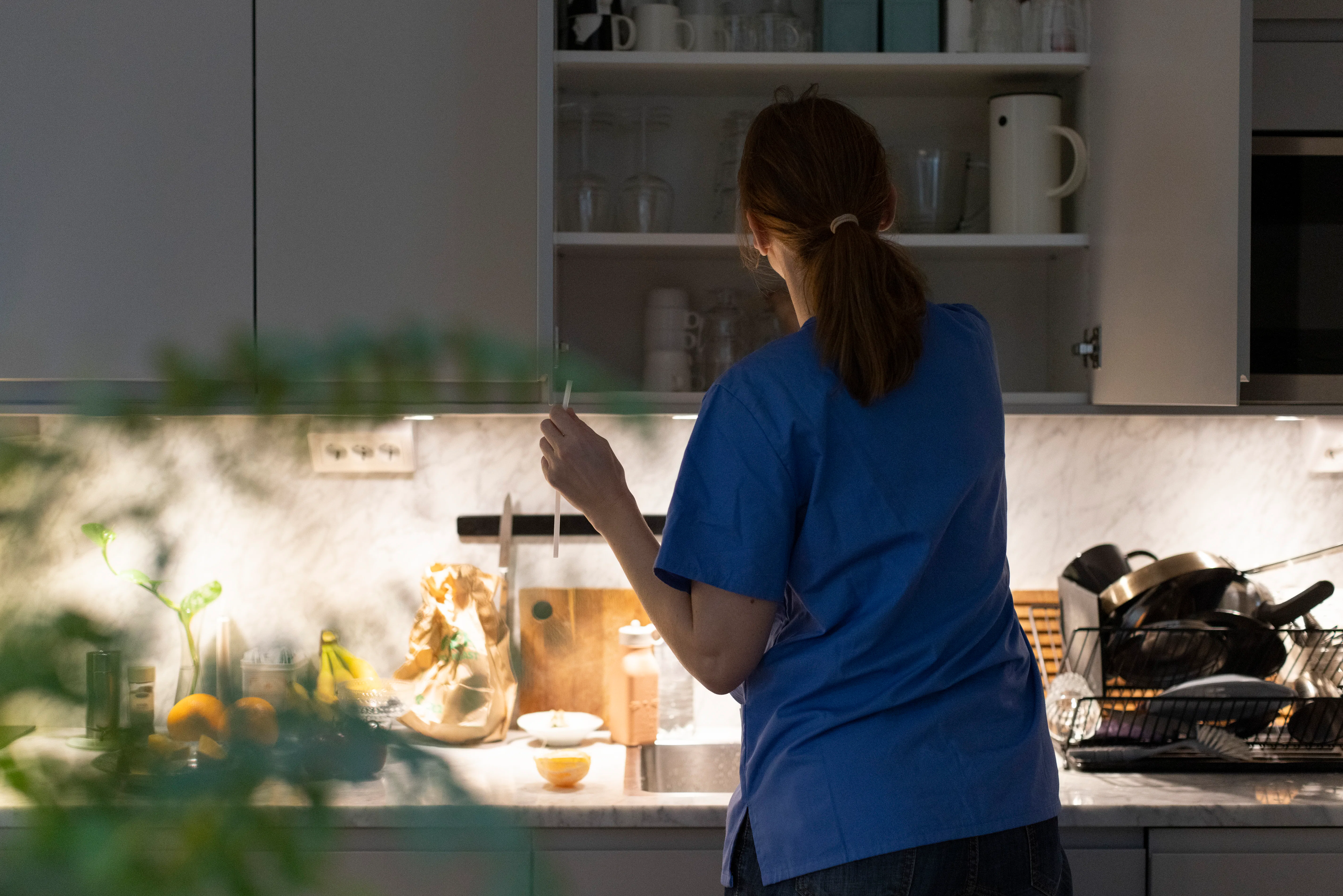 Person in blue scrub top, back to camera, reaching into an open kitchen cabinet above a marble count...