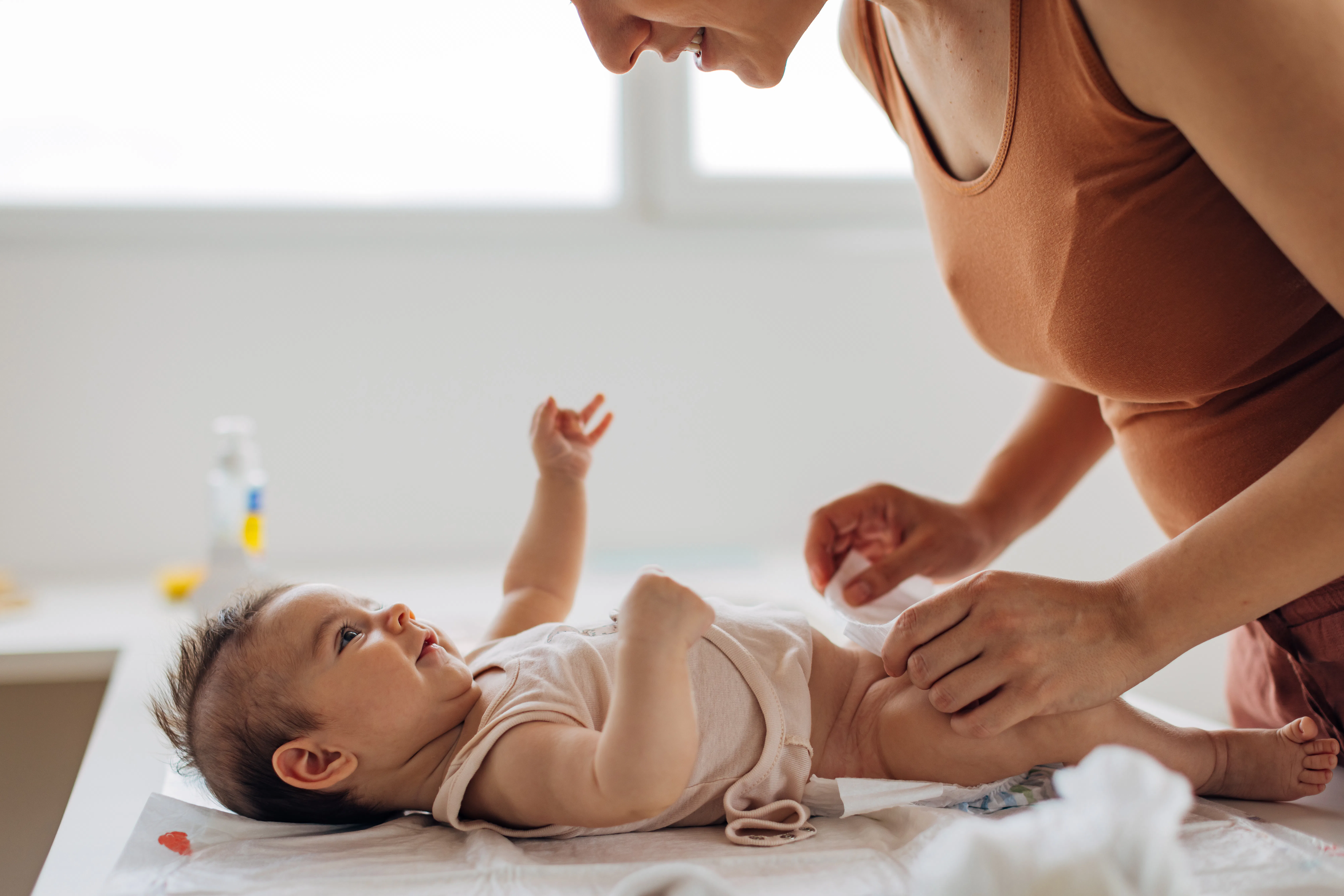 Parent leaning over changing table, smiling as they fasten a diaper on an infant lying on a pad, sof...
