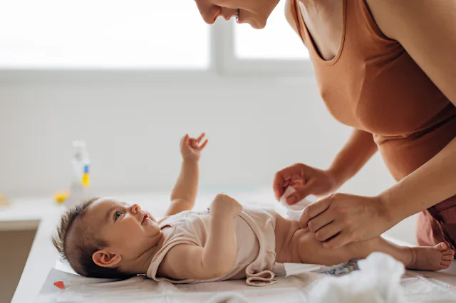 Parent leaning over changing table, smiling as they fasten a diaper on an infant lying on a pad, sof...