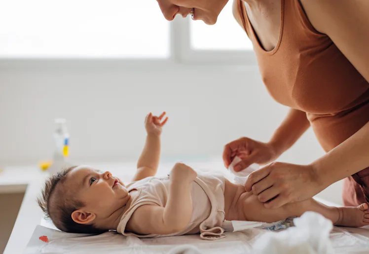 Parent leaning over changing table, smiling as they fasten a diaper on an infant lying on a pad, sof...