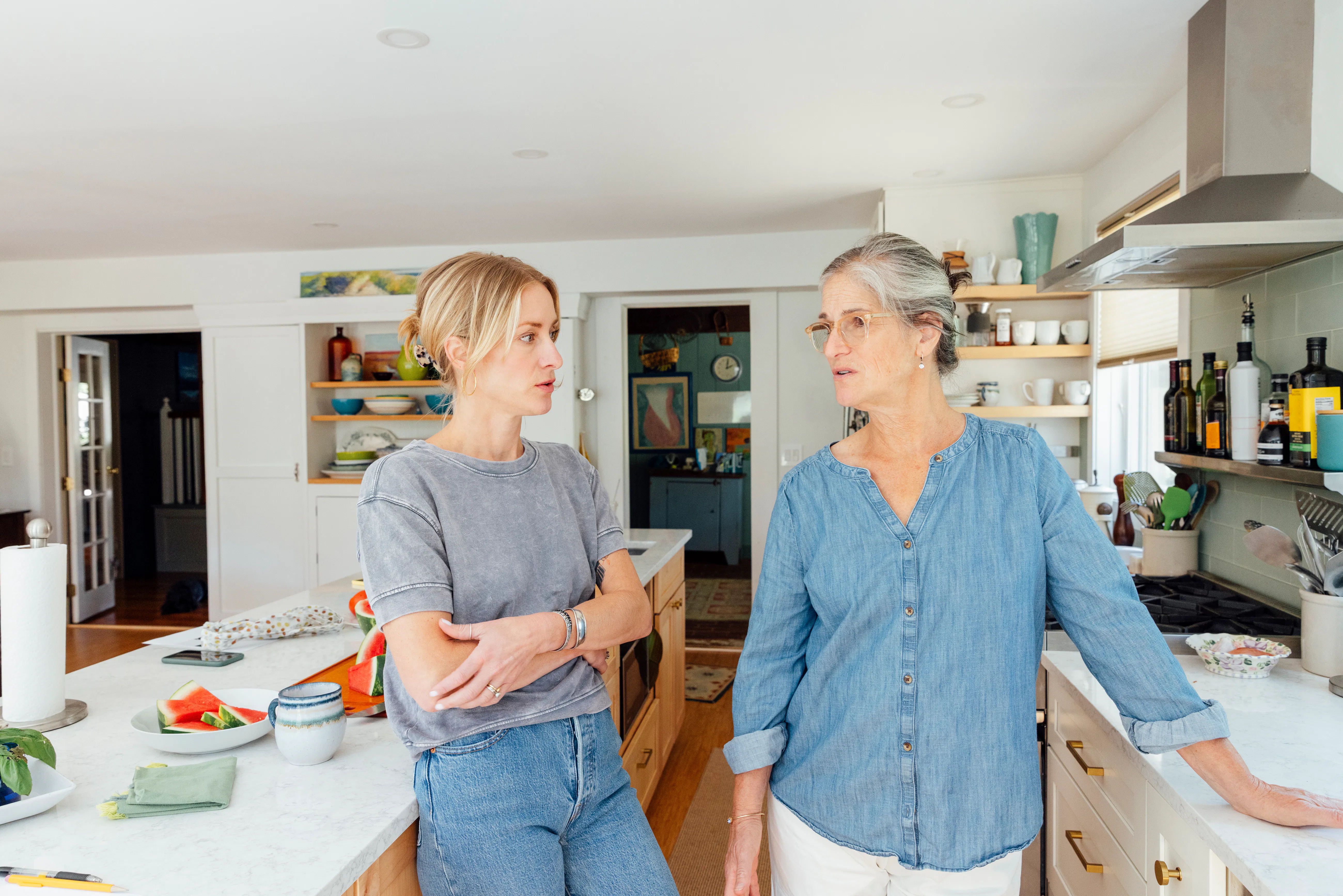 Two women stand in a bright kitchen, leaning on a marble island and countertops, engaged in conversa...