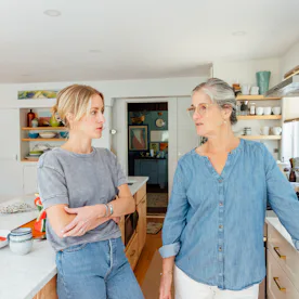 Two women stand in a bright kitchen, leaning on a marble island and countertops, engaged in conversa...