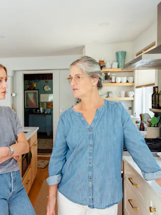 Two women stand in a bright kitchen, leaning on a marble island and countertops, engaged in conversa...