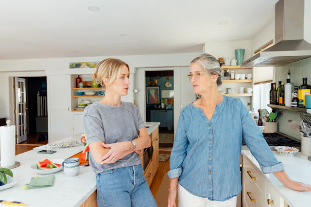 Two women stand in a bright kitchen, leaning on a marble island and countertops, engaged in conversa...