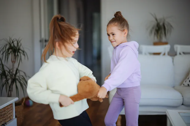 Two young girls in a living room tug on a teddy bear, one in a pale yellow sweatshirt and the other ...