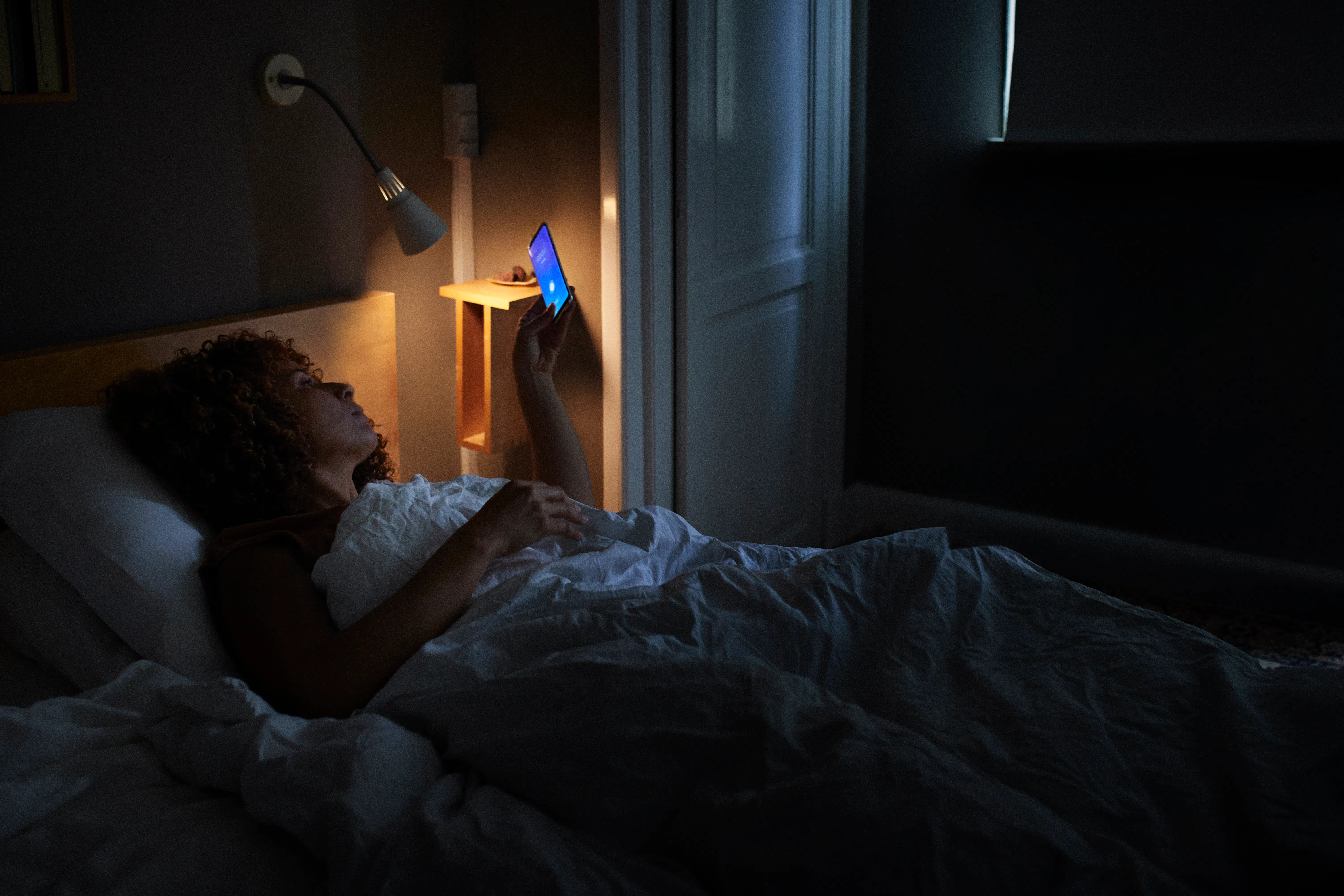 Person lying in bed at night, illuminated by a bedside lamp and the blue light of a smartphone held ...