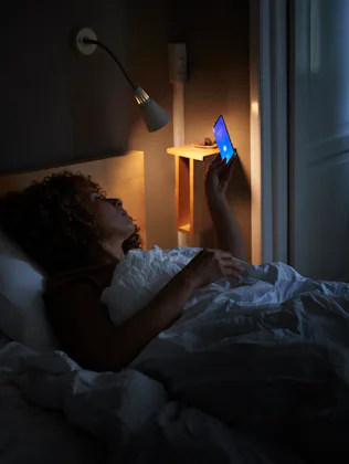 Person lying in bed at night, illuminated by a bedside lamp and the blue light of a smartphone held ...