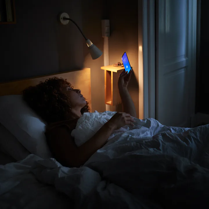 Person lying in bed at night, illuminated by a bedside lamp and the blue light of a smartphone held ...