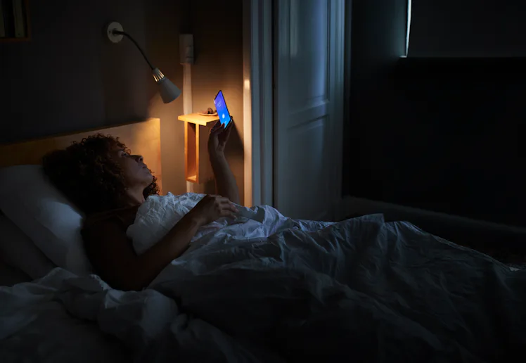 Person lying in bed at night, illuminated by a bedside lamp and the blue light of a smartphone held ...