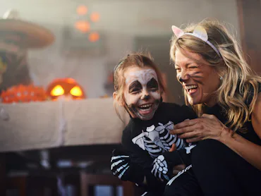 A smiling adult and child in Halloween costumes laugh together indoors; child wears skeleton face pa...