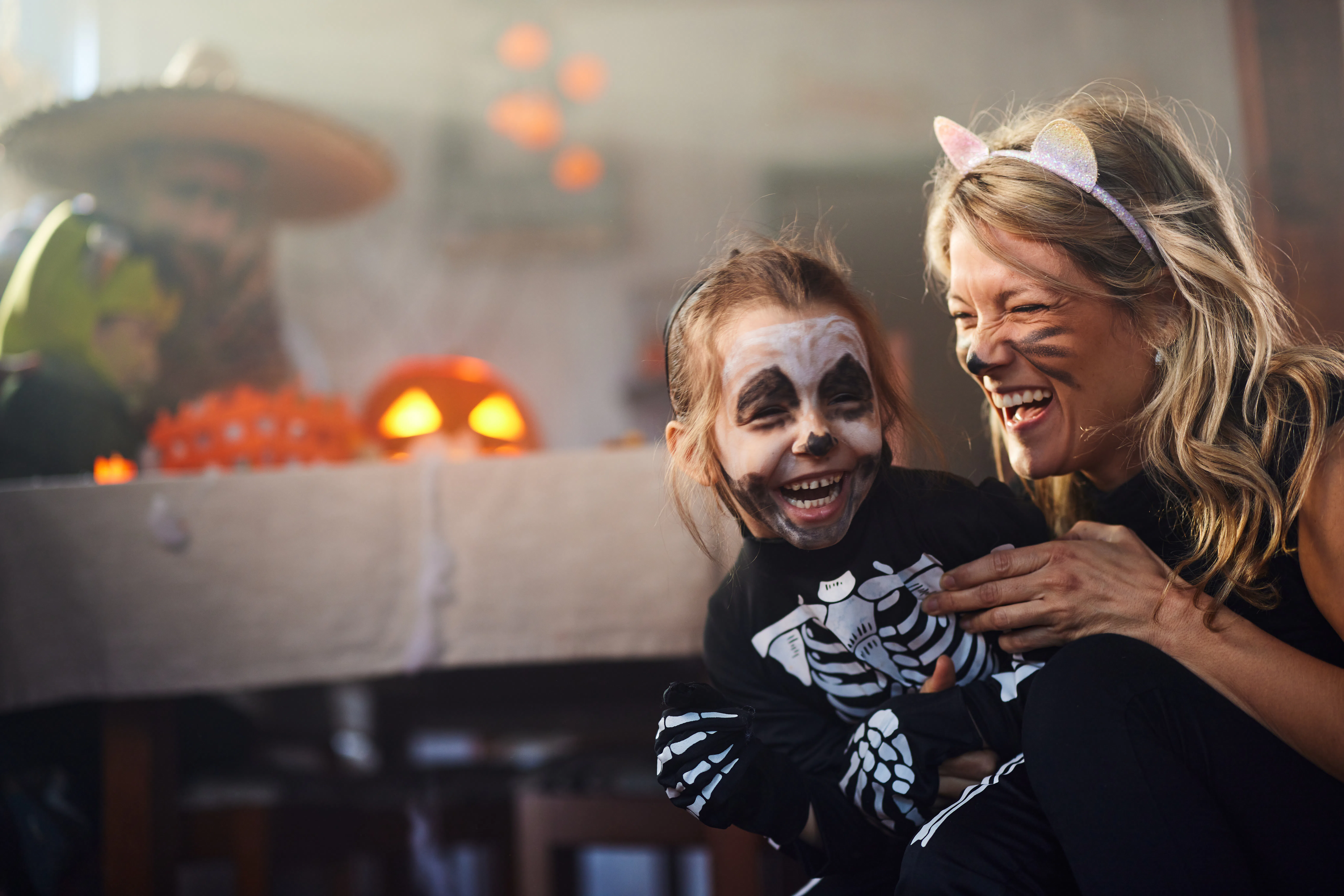 Woman wearing cat ears laughs while holding a child in skeleton face paint and costume in a dimly li...