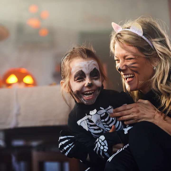 Woman wearing cat ears laughs while holding a child in skeleton face paint and costume in a dimly li...