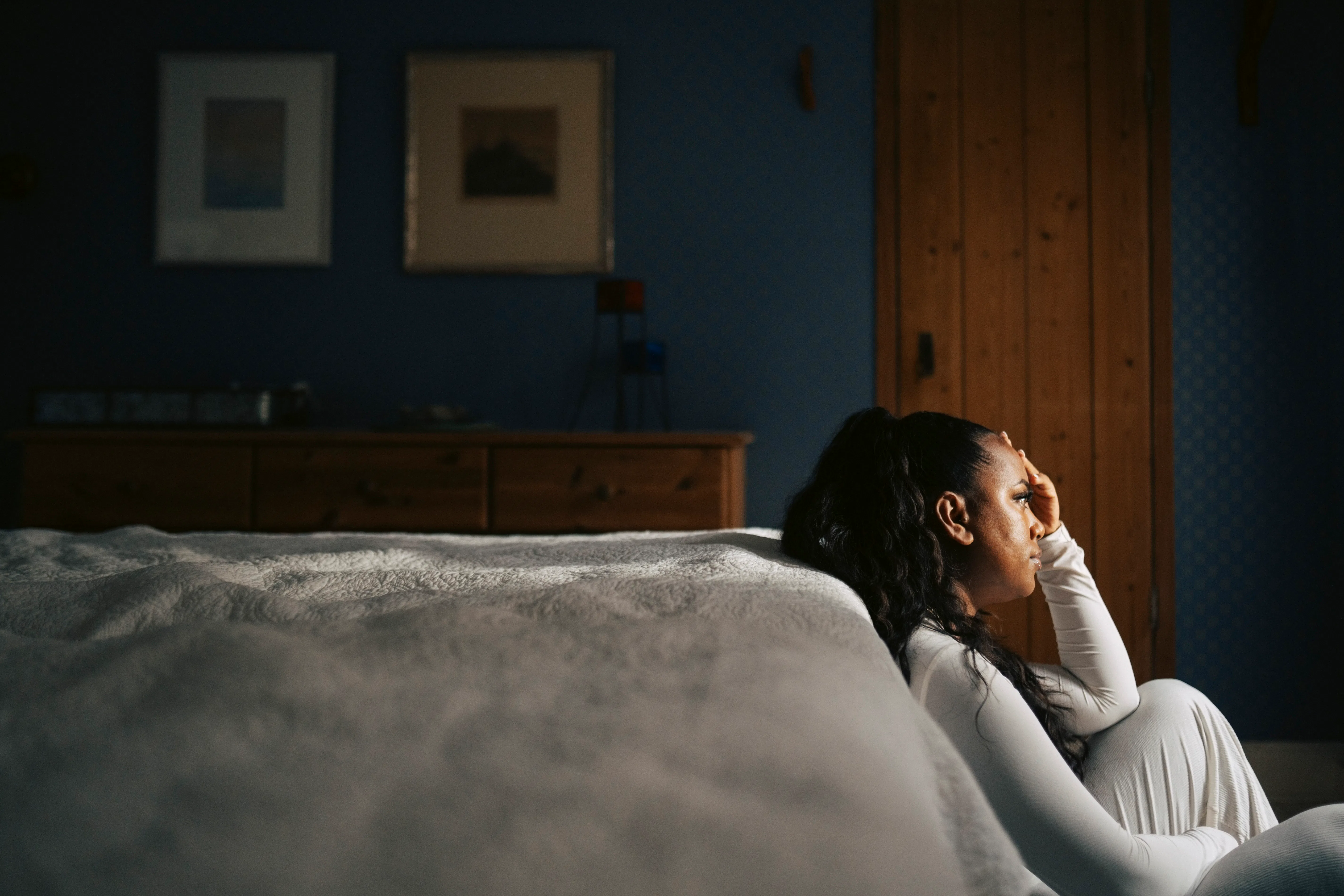 Woman in white sitting on bedroom floor beside bed, leaning against wall with hand to forehead, subd...