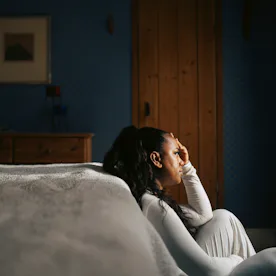 Woman in white sitting on bedroom floor beside bed, leaning against wall with hand to forehead, subd...
