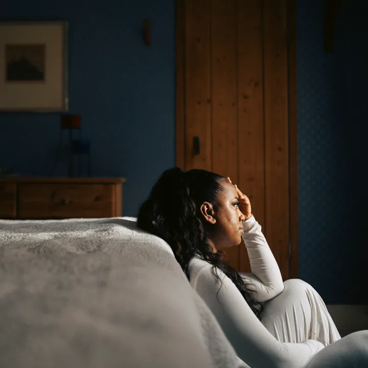 Woman in white sitting on bedroom floor beside bed, leaning against wall with hand to forehead, subd...
