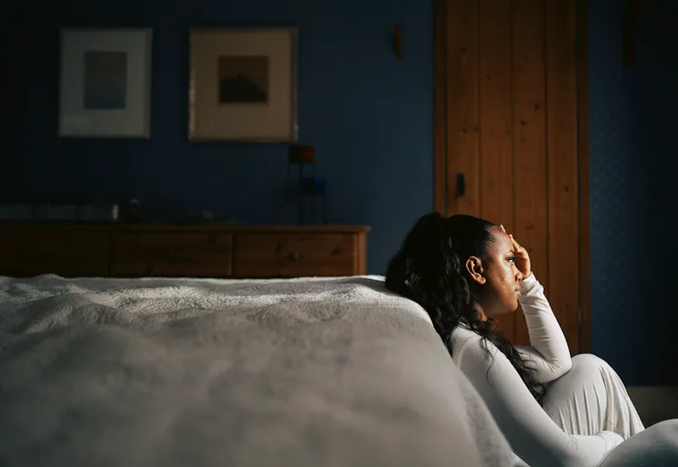 Woman in white sitting on bedroom floor beside bed, leaning against wall with hand to forehead, subd...