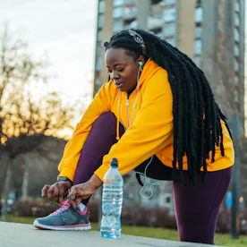 Person in a yellow hoodie and purple leggings tying a sneaker on an urban pathway at sunset, with ea...