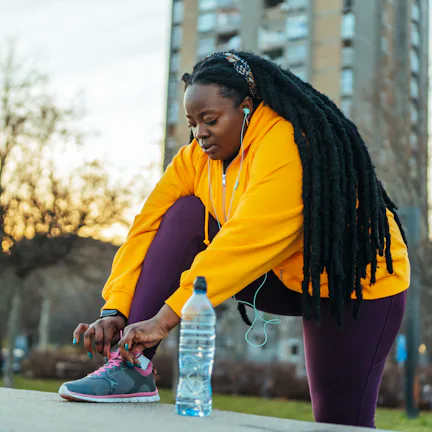 Person in a yellow hoodie and purple leggings tying a sneaker on an urban pathway at sunset, with ea...
