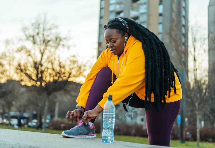 Person in a yellow hoodie and purple leggings tying a sneaker on an urban pathway at sunset, with ea...