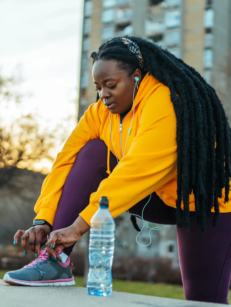 Person in a yellow hoodie and purple leggings tying a sneaker on an urban pathway at sunset, with ea...