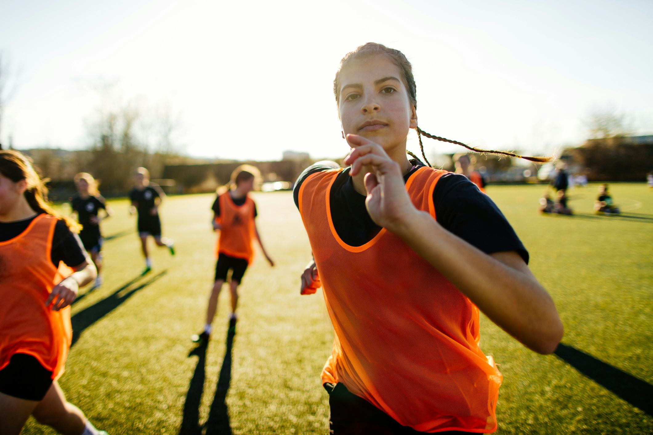 A young soccer player with braids, wearing an orange bib, runs towards the camera on a sunny field. ...