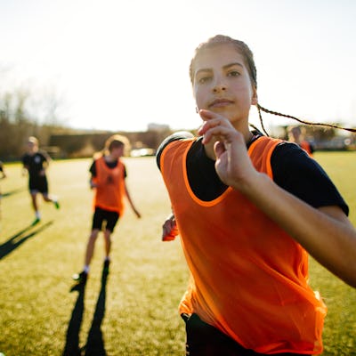 A young soccer player with braids, wearing an orange bib, runs towards the camera on a sunny field. Other players are visible in the background.