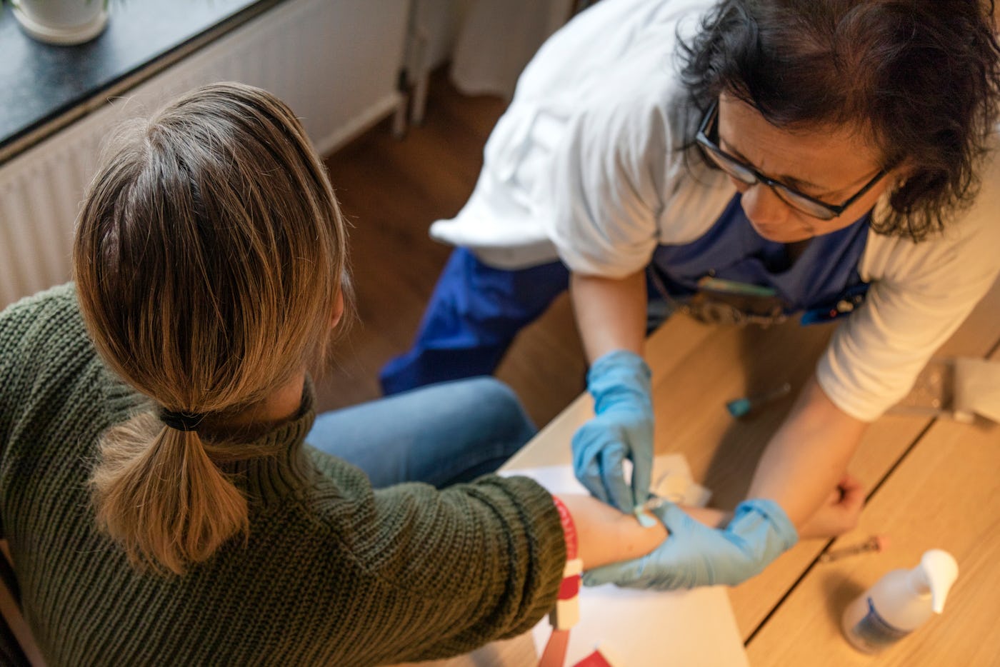 A healthcare worker in blue gloves is extracting blood from a person's arm using a needle and syringe. The setting appears to be a medical office.