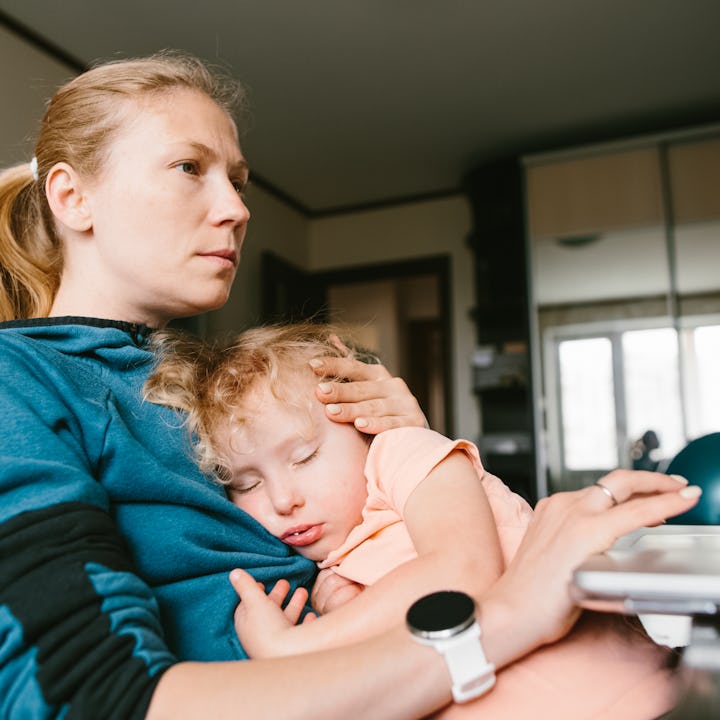 A woman in a blue hoodie is holding a sleeping child on her lap while sitting near a laptop in a roo...