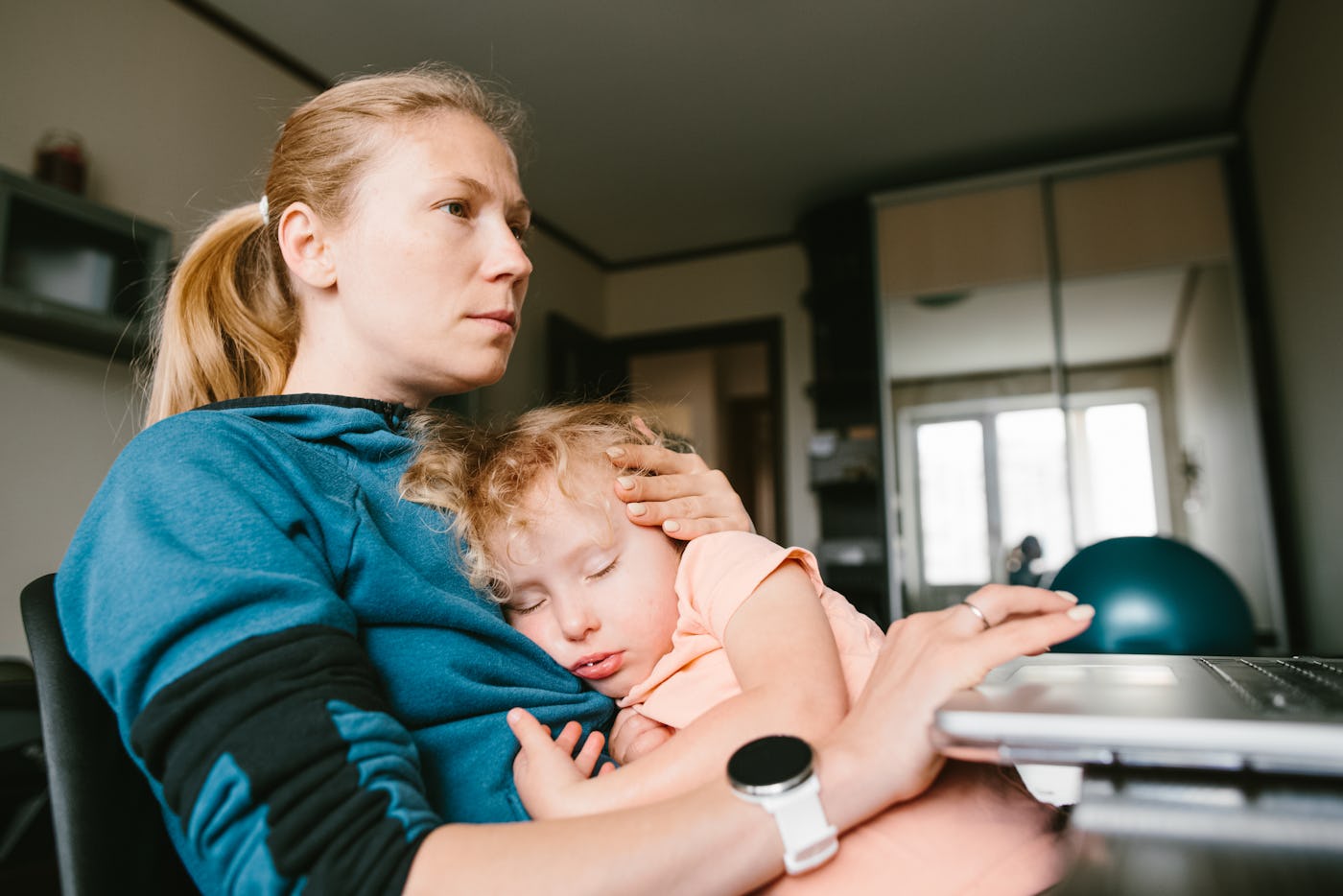 A woman in a blue hoodie is holding a sleeping child on her lap while sitting near a laptop in a room with a window and visible furniture.