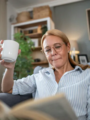 A person with glasses is sitting on a couch, holding a white mug and reading a book. The room has pl...