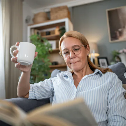 A person with glasses is sitting on a couch, holding a white mug and reading a book. The room has pl...