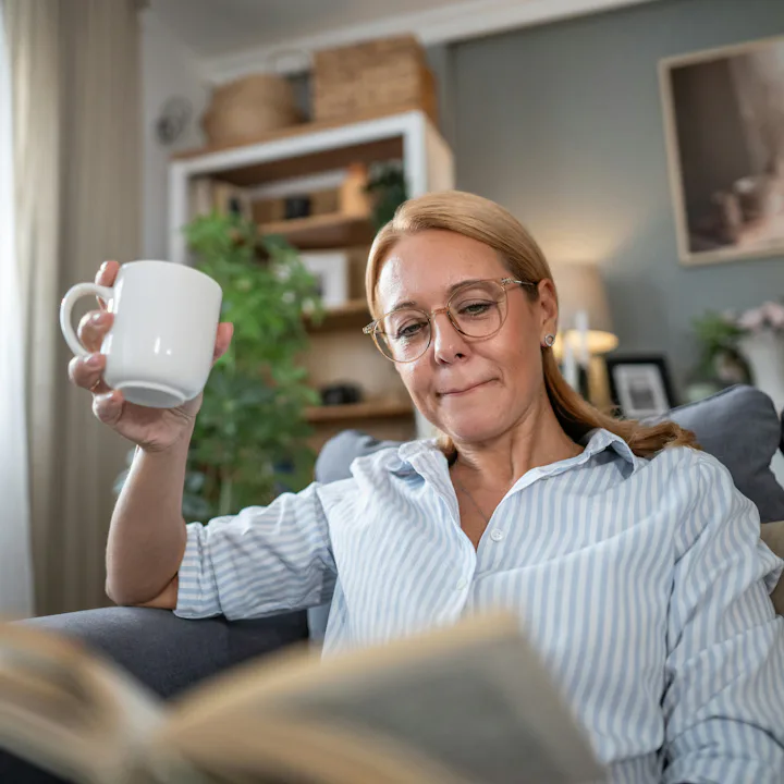 A person with glasses is sitting on a couch, holding a white mug and reading a book. The room has pl...