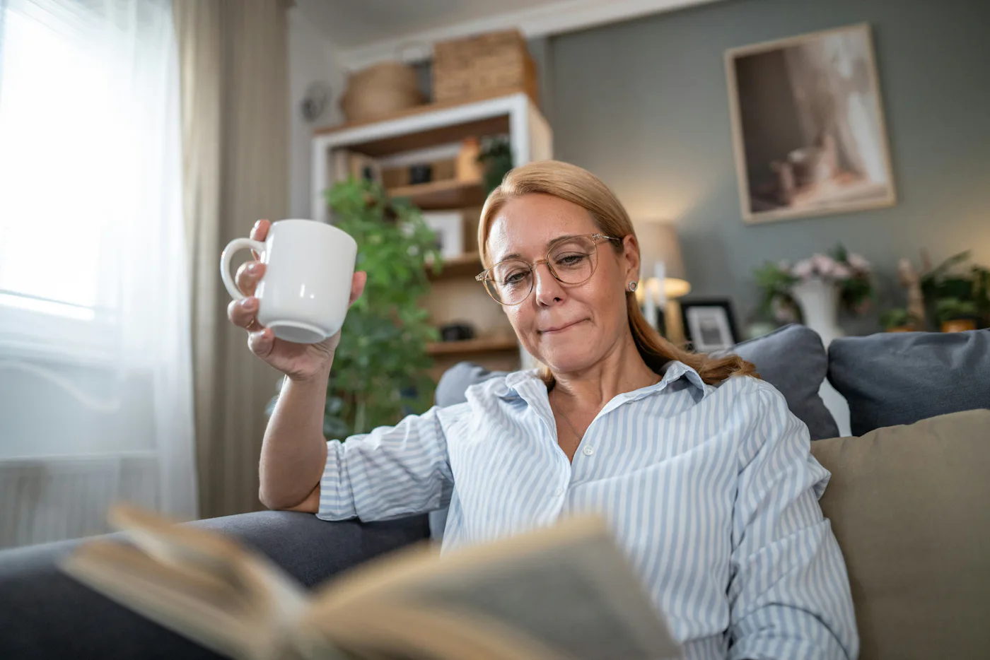 A person with glasses is sitting on a couch, holding a white mug and reading a book. The room has plants, a window, and framed art on the wall.