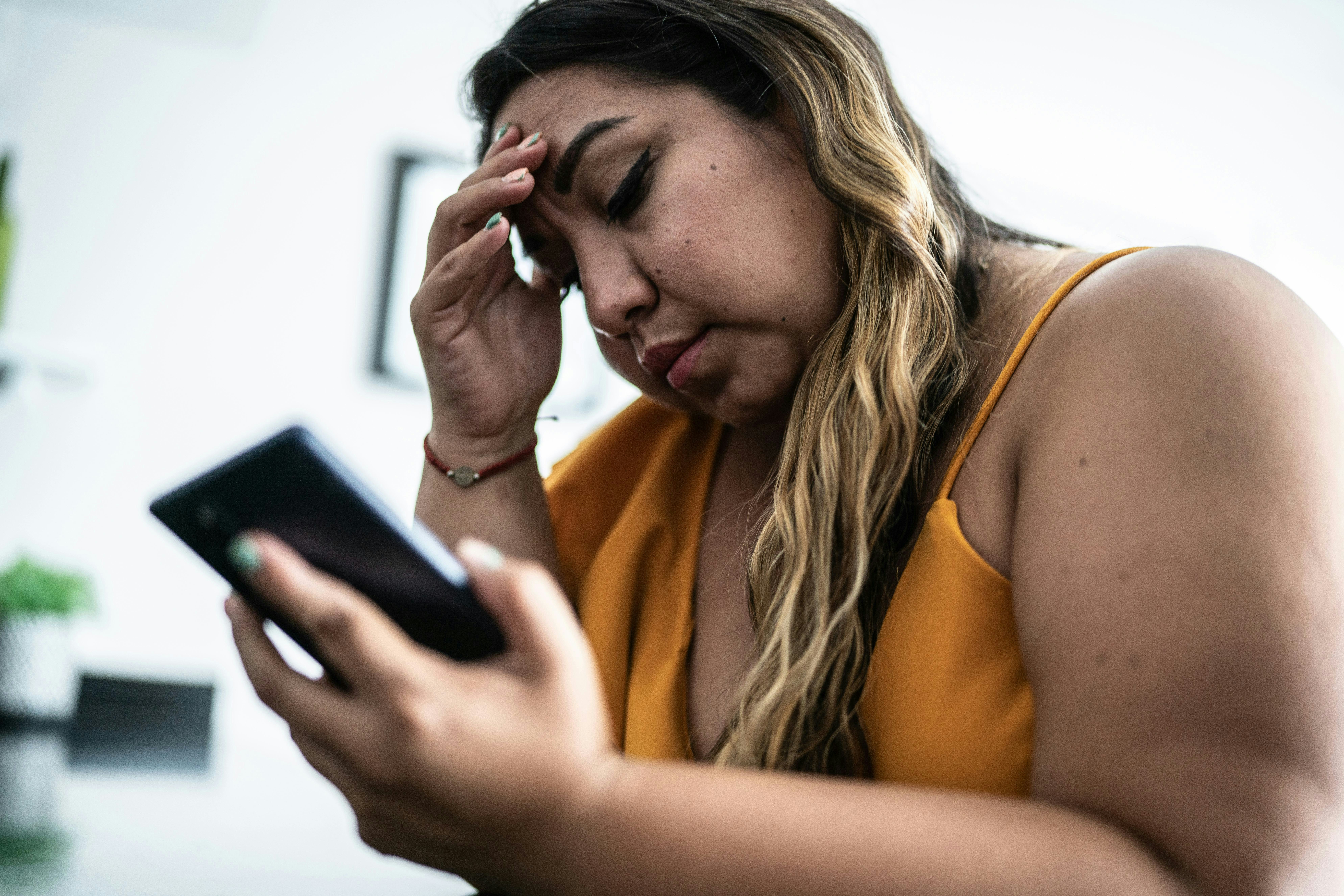 A woman with long hair is sitting, looking at her smartphone with a concerned expression, her hand o...