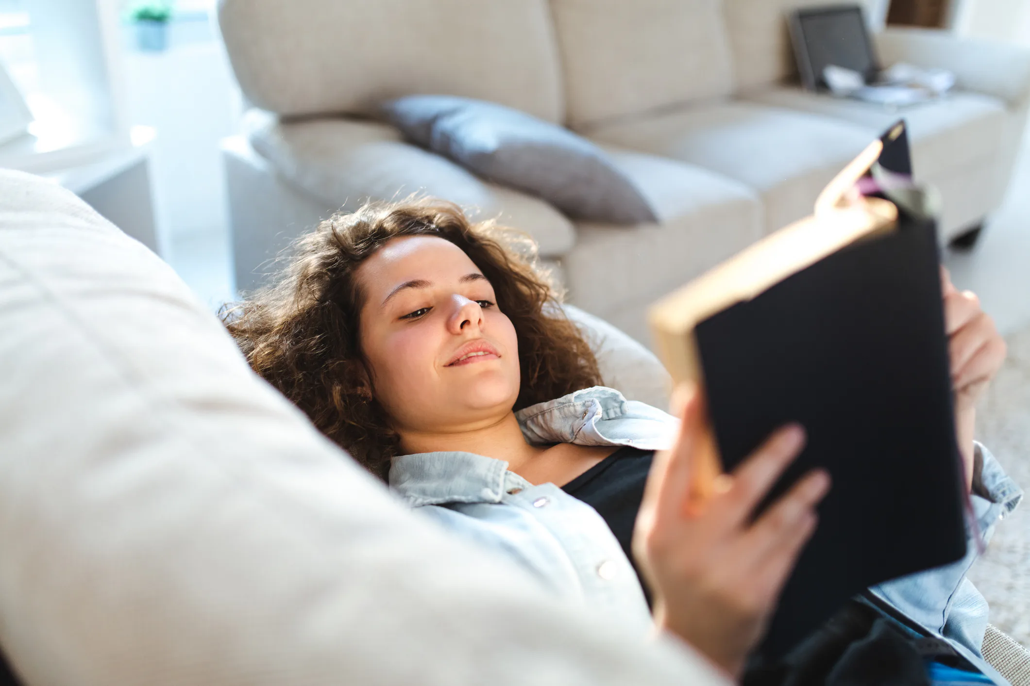 A person with curly hair is lying on a sofa, holding an open book. The background features a beige c...