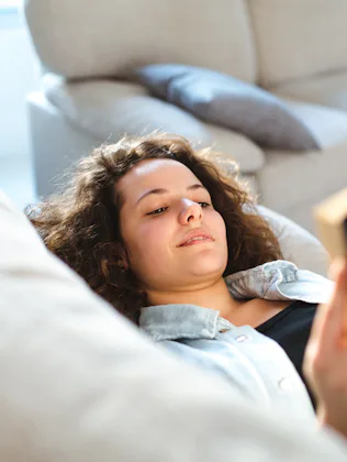 A person with curly hair is lying on a sofa, holding an open book. The background features a beige c...