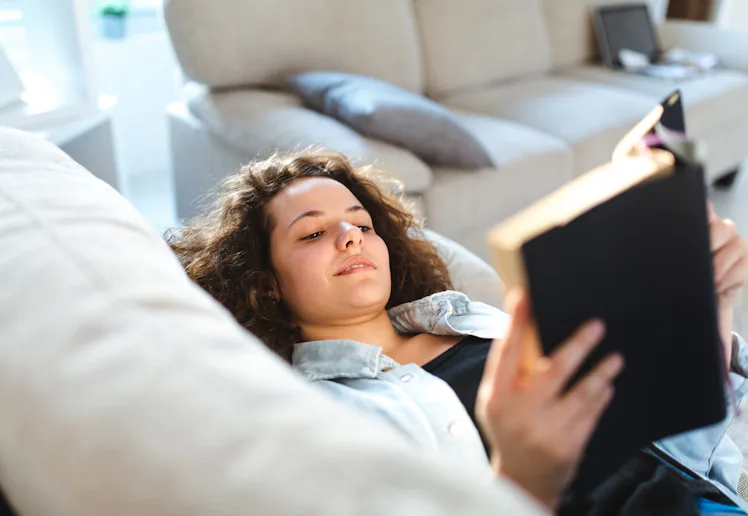 A person with curly hair is lying on a sofa, holding an open book. The background features a beige c...