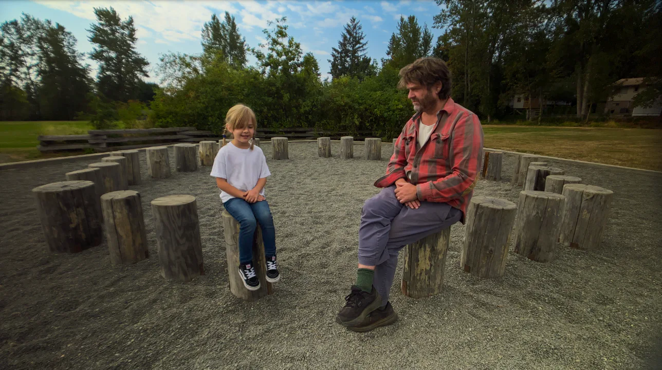 Zach Galifianakis sits on a tree stump talking to a child in a screen still from This Is A Gardening...