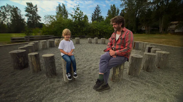 Zach Galifianakis sits on a tree stump talking to a child in a screen still from This Is A Gardening...